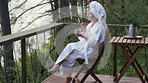 Young woman in a bathrobe and towel on the terrace on a background of mountains and forest. woman with a cup of tea and