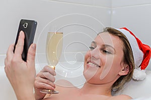 Young woman in the bathroom wearing a santa claus hat talks by video call on a smartphone and drinks champagne congratulating frie