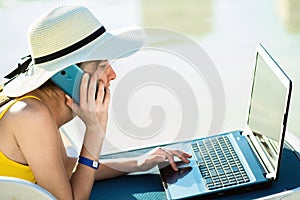 Young woman on beach chair at swimming pool working on computer laptop and talking on sell phone in summer resort. Remote work and