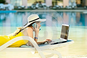 Young woman on beach chair at swimming pool working on computer laptop and talking on sell phone in summer resort. Remote work and