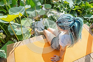 young woman with beautiful blue dreadlocks resting on yoga mat on lotus lake