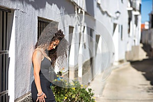 Young woman, beautiful, brunette, with curly hair, with a black dress and heels, alone and sad, leaning on a white house in a