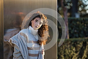 Young woman, beautiful, brunette with curly hair, sweater, coat and sunglasses, leaning on a metal wall, sensual and flirtatious