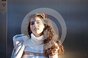 Young woman, beautiful, brunette with curly hair, sweater, coat and sunglasses, leaning on a metal wall, sensual and flirtatious