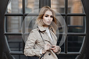 Young woman with beautiful eyes in a light spring coat with a fashionable black bag in a white t-shirt near brown door