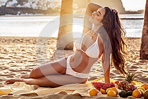 Young woman with beautiful fit tan body having picnic with fresh fruits at the empty tropical beach. Girl with long dark hair and