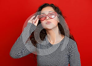 Young woman beautiful portrait flying kiss, posing on red background, long curly hair, sunglasses in heart shape, glamour concept