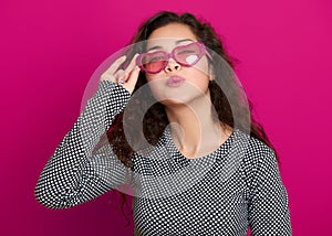 Young woman beautiful portrait make flying kiss, posing on pink background, long curly hair, sunglasses in heart shape, glamour co