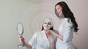 Young woman in beauty cosmetology salon with clay mask on face looks at the mirror and talks with female doctor