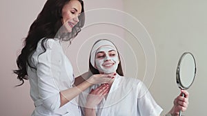 Young woman in beauty cosmetology salon with clay mask on face looks at the mirror and talks with female doctor