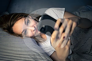 Young woman in bed holding a phone, tired and exhausted, blue light straining her eyes photo