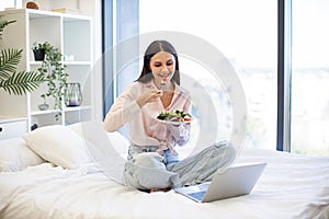 Young woman on bed at home and using modern laptop while eating healthy salad.