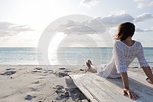 Young woman from behind sitting by the sea looks at the horizon at dawn in the wind, dressed in a white lace dress and white