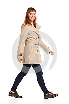 Young Woman In Beige Coat, Jeans And Brown Shoes Is Walking And Looking At Camera