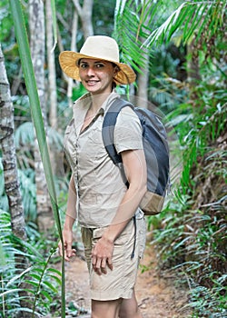 Young woman in beige shirt, shorts and straw haw, backpack on shoulders, smiling, jungle around