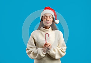 Young woman in beige sweater and Santa hat holding candy cane on light blue background. Celebrating Christmas