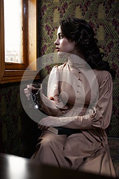 Young woman in beige vintage dress of early 20th century drinking tea in coupe of retro railway train