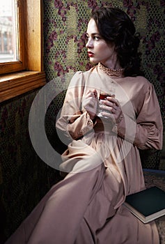 Young woman in beige vintage dress of early 20th century drinking tea in coupe of retro railway train