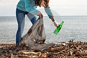 A young woman bends down to collect a dirty glass bottle. Cleaning and environmental protection