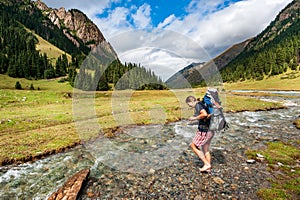 Young woman with big backpack barefoot crossing river. Shoeless female backapcker in river. Karakol valley on trail to Ala Kul