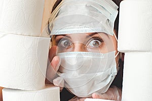 Young woman with big scared eyes in two medical virus protection face masks looks through stacks of toilet paper. Covid-19