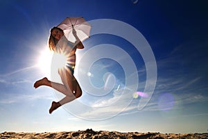 Young woman in bikini jumping over sand with umbrella in hands