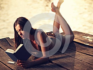 Young woman in bikini lying on jetty reading book