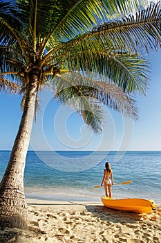 Young woman in bikini standing on a sandy beach with sea kayak and paddle