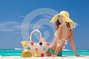 Young woman in bikini and straw hat and with colorful straw bag relaxing at white caribbean beach
