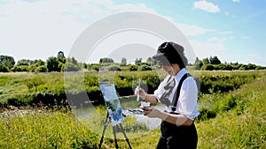 a young woman in a black cap stands on the Bank of a river and paints a landscape