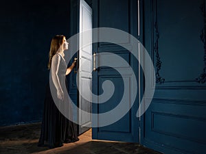 A young woman in a black dress opens a door from which light is pouring.