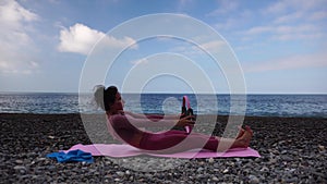 A young woman with black hair doing Pilates with the ring on the yoga mat near the sea on the pebble beach. Female