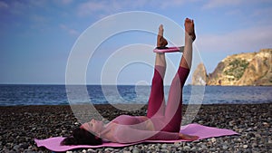 A young woman with black hair doing Pilates with the ring on the yoga mat near the sea on the pebble beach. Female