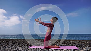 A young woman with black hair doing Pilates with the ring on the yoga mat near the sea on the pebble beach. Female