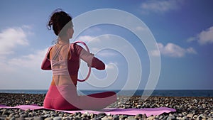 A young woman with black hair doing Pilates with the ring on the yoga mat near the sea on the pebble beach. Female