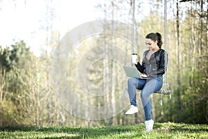 Young woman in a black jacked with coffee cup and laptop working outside in a park. Remote work. Digital work.