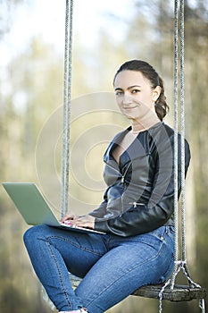 Young woman in a black jacked with coffee cup and laptop working outside in a park. Remote work. Digital work.