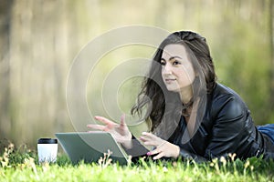 Young woman in a black jacked with coffee cup and laptop working outside in a park. Remote work. Digital work.