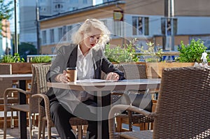 Young woman in a black jacket and glasses sitting at a table in street cafe, looking through documents. Business activity,