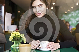 Young woman in black jersey look to camera, drink coffee and having rest in cafe near window.