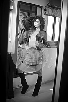 Young woman in black leather jacket and gray short tutu skirt looking into a large mirror. Beautiful curly dark hair girl posing