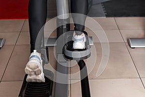 Young woman in black leggings in white sneakers walks on a stepper simulator in the gym. Girl does cardio to burn calories.