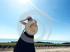 Young woman in black long dress and wide brimmed straw hat stands in graceful pose and looks at vineyard. Slender girl stands back photo
