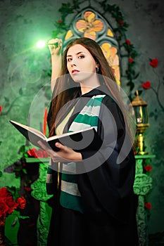 A young woman in a black mantle with a striped scarf around her neck casts a spell from a book and conjures with a wand of magic.