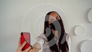 Young woman in black medical mask taking selfie on smartphone on background of white wall. Portrait of female spending time weeken