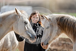 Young woman in black riding jacket standing near group of white Arabian horses smiling happy, one on each side, closeup detail