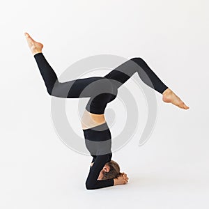 A young woman in black sportswear, performs a variation of the shirshasana exercise, trains in the studio on a light background