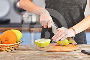 A young woman in a black t-shirt cuts green apple on a light wooden board in a kitchen