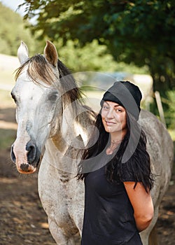 Young woman in black t shirt and hat, standing next to her white Arabian horse, blurred yard with trees background