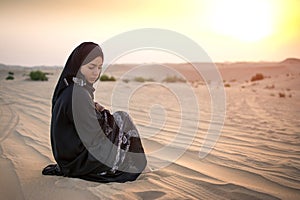 Young woman in black traditional clothes sitting on sand against sunset over desert